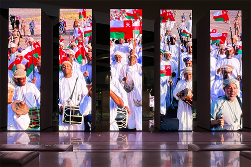 Series of vertical screens displaying footage of Omani men in traditional dress carrying flags and drums, part of a museum exhibition display.