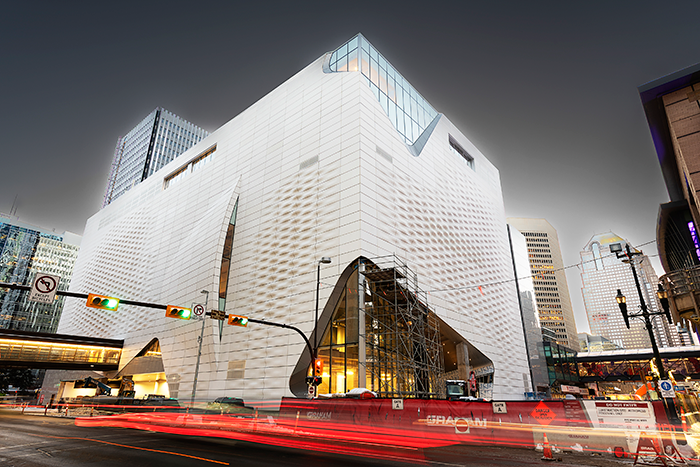 The Glenbow Art Museum under construction at night, its angular white facade rising above a busy Calgary intersection with light trails from passing traffic.