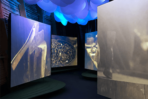 Curved video screens displaying close-up footage of hands and craft materials in the Irish Pavilion, set beneath a sculptural blue cloud installation.
