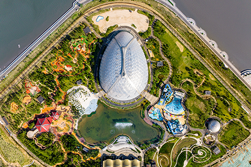 Aerial view of the Eden Qingdao park, with a large dome-shaped greenhouse at the centre surrounded by themed gardens, water features, and a coastal waterfront setting.