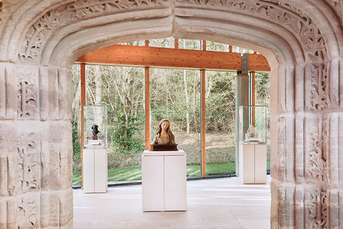 Stone-carved archway leading into a gallery at the Burrell Collection, with sculptures displayed on white plinths against floor-to-ceiling windows overlooking woodland.