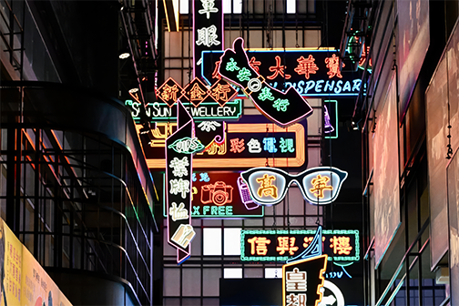 Illuminated neon signage in Chinese and English, including a giant pair of glasses and shop signs, in a recreated Hong Kong street scene.