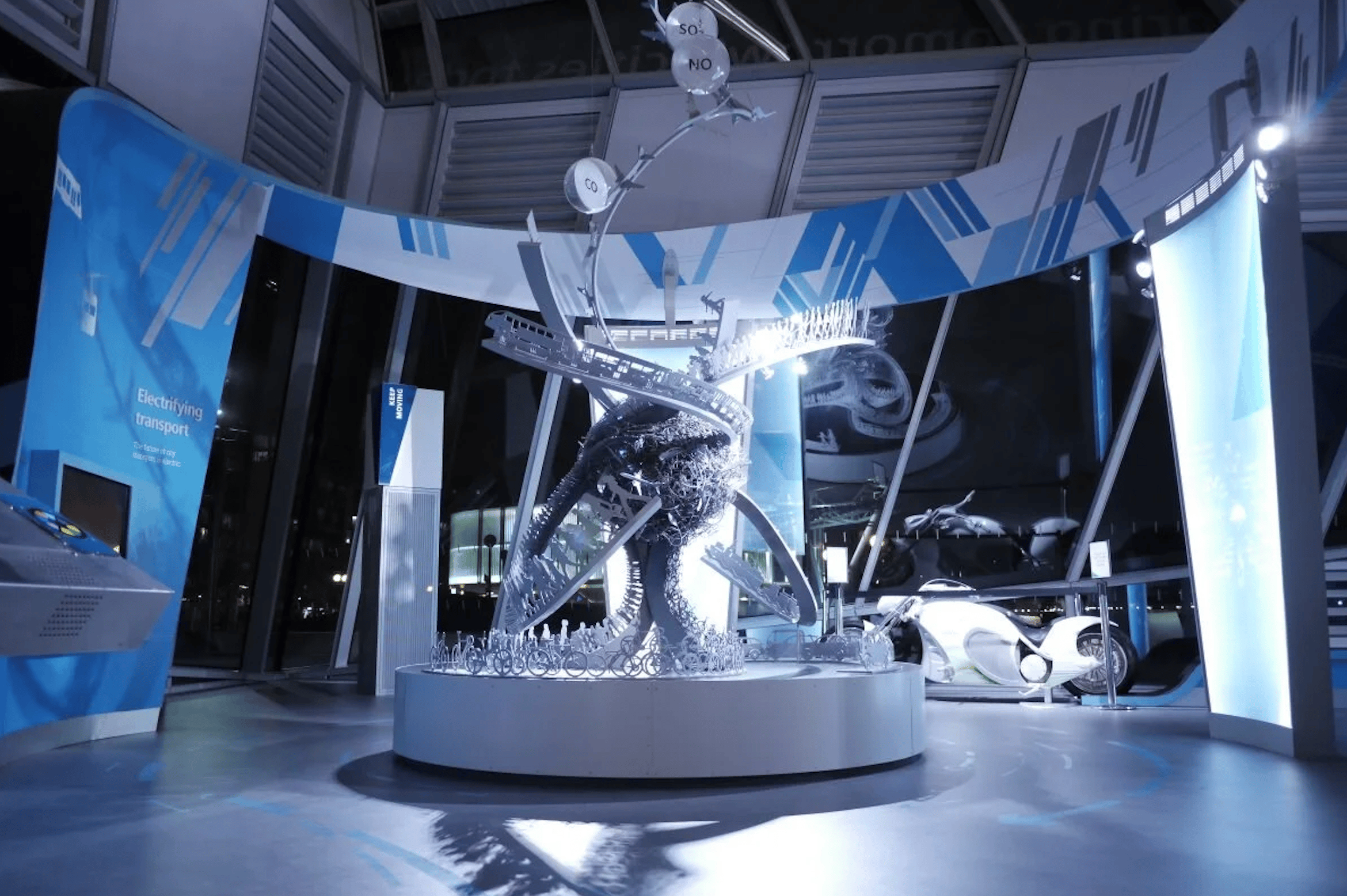 Sculptural installation of intertwined transport vehicles at The Crystal's 'Electrifying Transport' exhibition, with a futuristic electric vehicle displayed nearby.