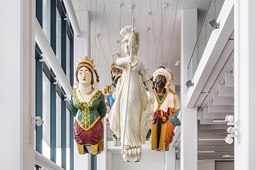 Collection of painted ship figureheads suspended in The Box museum's atrium, depicting historical and royal figures above a white staircase.