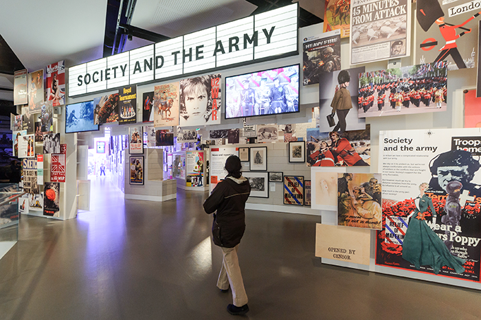 Visitor viewing the 'Society and the Army' gallery at the National Army Museum, with walls covered in historical posters, photographs, and display screens.